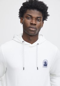 Sweat-shirt à capuche blanche avec une poche kangourou à l'avant et des détails en logo bleu. Il comprend des cordons de serrage réglables avec des embouts noirs et un tissu texturé.