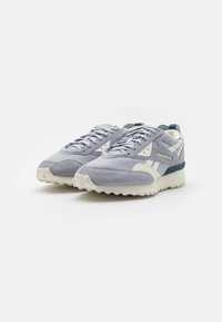 Šedé semišové tenisky s bílými akcenty, texturovanou gumovou podrážkou a kontrastním tmavě modrým zadním dílem. Mají tkaničky a logo na boku.