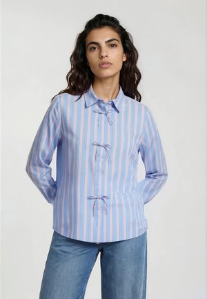 Frau trägt ein blaues Langarmshirt mit vertikalen pinken und weißen Streifen und gebundenen Schleifen vorne, kombiniert mit blauen Jeans.