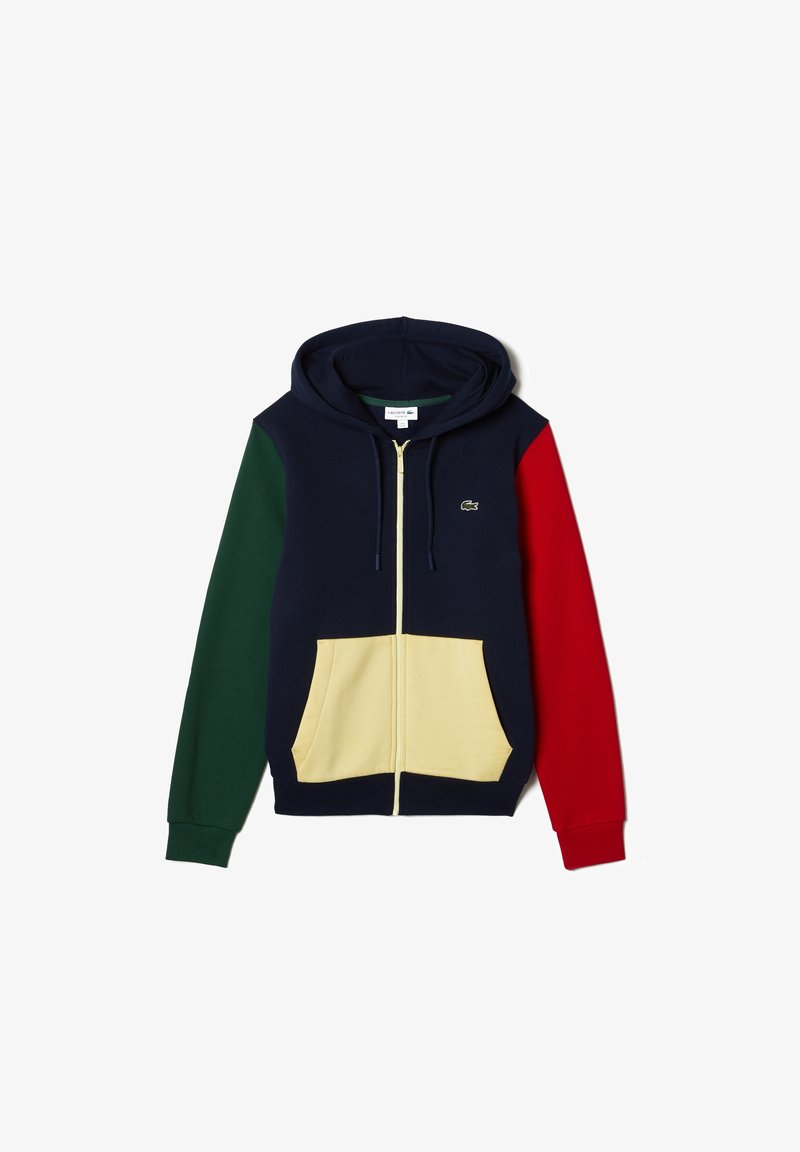 Mörkblå zip-hoodie med mörkgrön vänsterärm, röd högerärm, beige frontfickor och en liten grön krokodil-logotyp på bröstet.