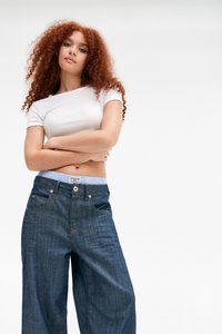 Γυναίκα με σγουρά κόκκινα μαλλιά, φορώντας ένα λευκό cropped top και χαλαρούς σκούρους μπλε τζιν, σταθεί με τα χέρια σταυρωμένα μπροστά σε άσπρο φόντο.