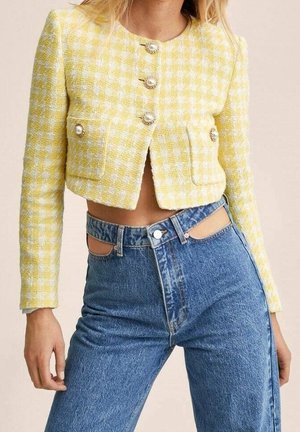 Vrouw draagt een geel-wit geblokt cropped jasje met gouden knopen en een blauwe hoge taille spijkerbroek met uitsnijdingen aan de taille.