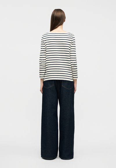 Άτομο με μακριά καστανά μαλλιά που φοράει λευκή και ναυτική μπλούζα με ρίγες μακριάς μανίκας και φαρδιά μπλε τζιν παντελόνια, γυρισμένο πλάτη.