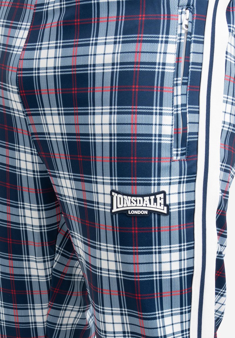 Marineblaue Karottenhosen mit roten und weißen Akzenten, ausgestattet mit einem Lonsdale London Logo-Patch und seitlichen Reißverschlusstaschen. Glatte Stoffoberfläche.