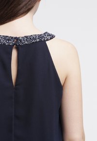 Marinblus utan ärmar med en dekorativ pärlhalskrage och knappstängning bak. Slät tyg, avslappnad passform, elegant design.