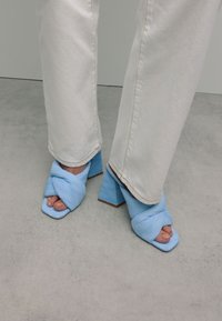 Lichtblauwe hakken sandalen met een gedraaide stofontwerp en vierkante neus. De hakken zijn dik en gestructureerd, gecombineerd met witte rechte pijpen.