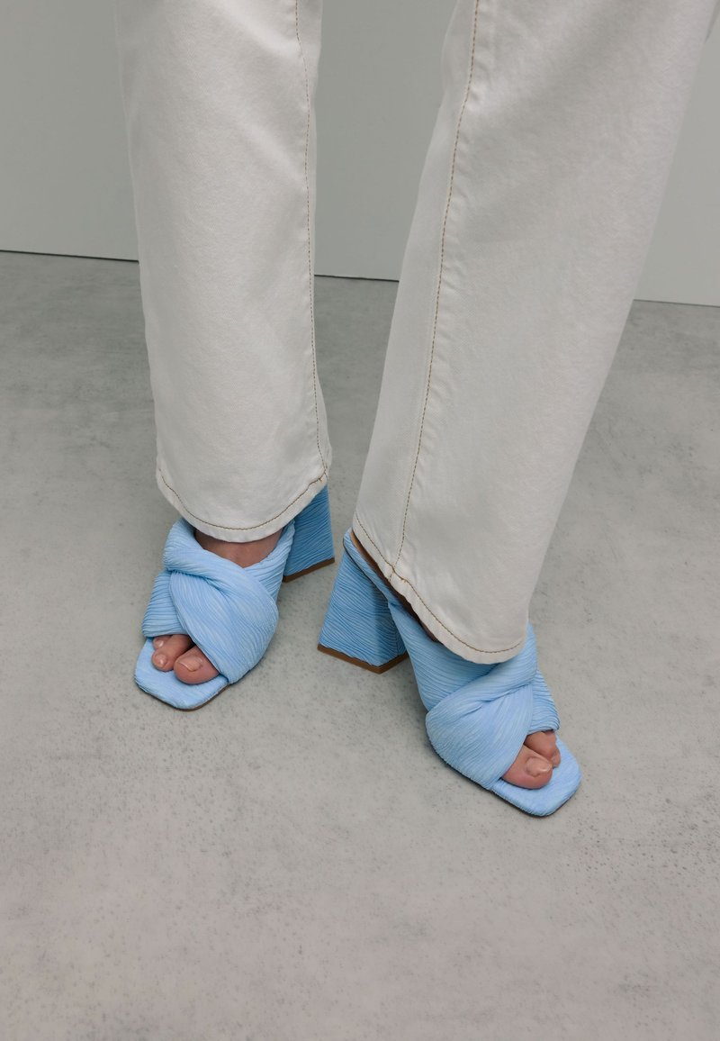 Lichtblauwe hakken sandalen met een gedraaide stofontwerp en vierkante neus. De hakken zijn dik en gestructureerd, gecombineerd met witte rechte pijpen.