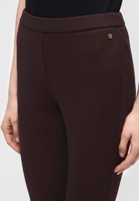 Gros plan d'une personne portant un pantalon marron foncé ajusté et un haut noir, montrant la taille et les mains posées à côté des jambes.