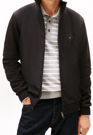 Mann trägt schwarze Reißverschlussjacke über grau-weiß gestreiftem Polohemd mit dunklen Jeans, Hände in den Taschen, vor weißem Hintergrund.