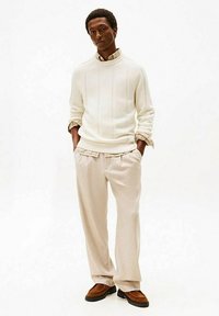 Maglione bianco a maglia con trama a coste, pantaloni beige plissettati, scarpe in camoscio marrone e colletto di camicia a quadri visibile sotto il maglione.