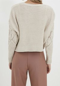 Světlý béžový pletený svetr s texturovaným vzorem a žebrovaným lemem. Rukávy zdobené copánkovým vzorem. Kombinováno s béžovými kalhotami.