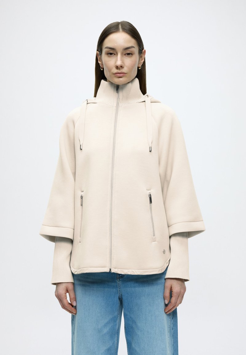 Μπεζ μπουφάν με φερμουάρ, ψηλό γιακά, φαρδιά μανίκια και τσέπες μπροστά με φερμουάρ. Κατασκευασμένο από απαλό ύφασμα με ομαλή υφή.