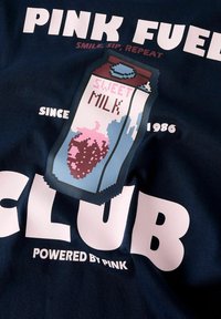 Granatowa koszulka z tkaniny z grafiką w stylu pixel art przedstawiającą karton mleka z napisem "SŁODKIE MLEKO" oraz dużym tekstem "PINK FUELED CLUB" i "POWERED BY PINK."