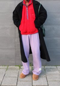 Pullover rojo de forro polar con media cremallera, abrigo largo negro, pantalones de chándal lavanda y zapatos tipo slip-on color tostado, de pie sobre un pavimento gris con fondo texturizado.