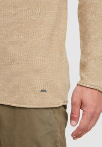 Beige Strickpullover mit einem strukturierten Muster, featuring einen abgerundeten Saum und einem Etikett am unteren Ende. Die Hand des Models ruht auf einer olivgrünen Hose.