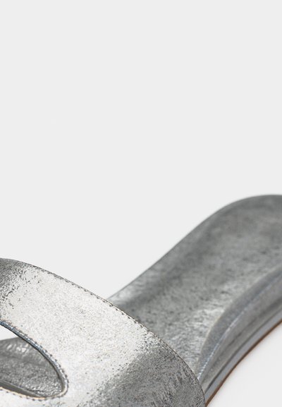 Ανοιχτό πέδιλο σε ασημί μεταλλικό χρώμα με φαρδιά λουρί και επίπεδη σόλα σε λευκό φόντο.