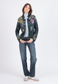 Cardigan floreale verde con bottoni davanti, caratterizzato da ricami floreali gialli e rosa. Abbinato a un dolcevita grigio a coste e jeans blu scuro.