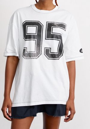 Biała oversize koszulka z czarną, błyszczącą cyfrą „95” na przodzie, z krótkimi rękawami i okrągłym dekoltem. Czarna naszywka Nike na rękawie.