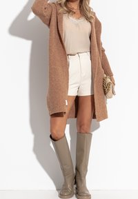 Langer, gestrickter Cardigan in Beige, über einem hellbeigen Oberteil und weißen Shorts getragen. Kombiniert mit knielangen, glänzenden beigen Stiefeln und einer gestepphten Handtasche.