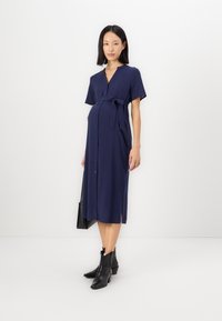 Ναυτικό μπλε μητρότητα φόρεμα με κουμπιά μπροστά, κοντό μανίκι και ζώνη στη μέση. Συνδυασμένο με μαύρες αστραγάλες μπότες και μαύρη τσάντα χειρός.