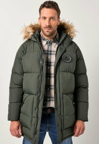 Homme portant un manteau d'hiver rembourré vert avec une capuche en fourrure, une chemise à carreaux et un jean bleu, faisant face à l'avant sur un fond clair.
