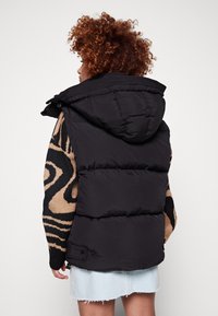 Svart puffer-väst med huva, med mönstrade stickade ärmar i beige och svart, samt en något cropped passform med två sidofickor.