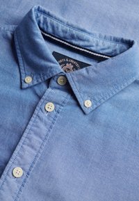 Chemise bleu clair à boutons blancs, col plié et étiquette visible à l'intérieur du col.