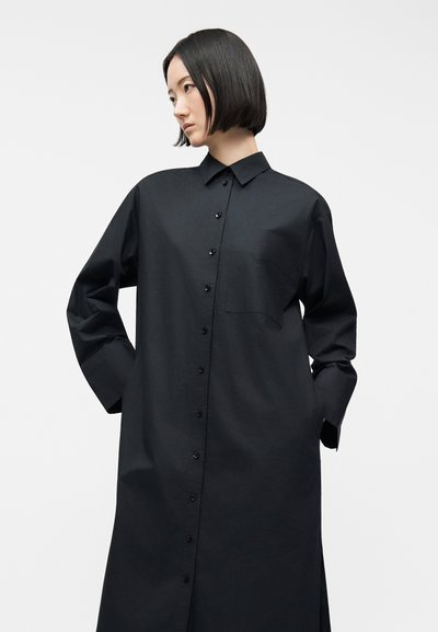 Γυναίκα με κοντά μαύρα μαλλιά, φορώντας μακριά μαύρη πουκαμίσα-φόρεμα με γιακά και κουμπιά, με τα χέρια στις τσέπες, μπροστά από απλό φωτεινό φόντο.