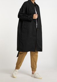 Abrigo negro de gran tamaño con cierre de botón, dos bolsillos delanteros y cuello entallado, combinado con pantalones jogger color beige y zapatillas blancas.