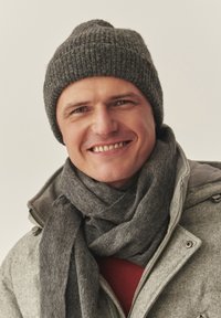 Szara dzianinowa czapka typu beanie, dopasowany szary szalik oraz jasnoszara kurtka na suwak. Tło jest neutralne; osoba na zdjęciu lekko się uśmiecha.