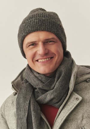 Harmaa neulottu pipo, siihen sopiva harmaa huivi ja vaaleanharmaa vetoketjullinen takki. Tausta on neutraali; henkilöllä on kevyt hymy.