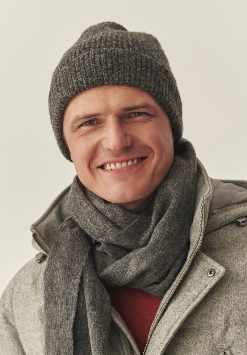 Szara dzianinowa czapka typu beanie, dopasowany szary szalik oraz jasnoszara kurtka na suwak. Tło jest neutralne; osoba na zdjęciu lekko się uśmiecha.