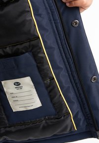 Interno della giacca navale con fodera trapuntata nera, bordo giallo e un'etichetta in tessuto che riporta "Petrol Industries." Presenta una chiusura a scatto e cerniera.