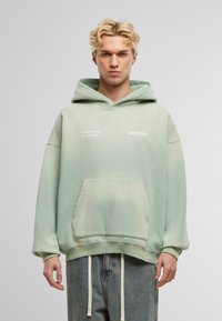 Sudadera oversized verde menta con un bolsillo frontal, acentos de texto en blanco y puños de canalé. Combinada con jeans de lavado claro y detalles de cordón.
