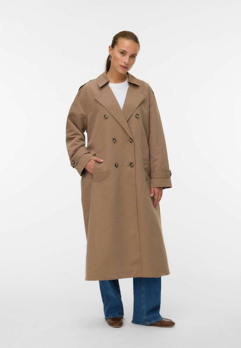 Khaki trench coat z dvojitimi gumbi, stranskimi žepi in ovratnikom z režami, v kombinaciji z modrimi kavbojkami in rjavimi balerinkami.
