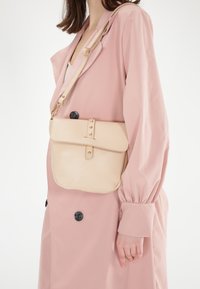 Beige tekstureret crossbody taske med klap, gyldne hardware detaljer og en justerbar rem, båret mod en lyserød frakke med sorte knapper.