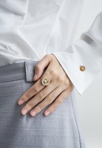 Χρυσό δαχτυλίδι με σχέδιο σε σχήμα C, διακοσμημένο με διαυγείς κρυστάλλους και υφή στην ταινία, φορεμένο σε χέρι πάνω σε ανοιχτό γκρι ύφασμα.