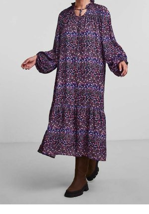 Frau trägt ein langes lila Blumenmuster-Kleid mit Puffärmeln und braunen kniehohen Stiefeln, steht auf einem grauen Boden.