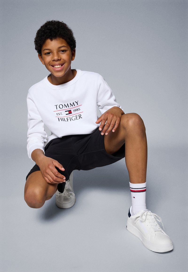 Fehér pulóver "TOMMY HILFIGER" logóval, fekete rövidnadrág, fehér sportcipő, valamint fehér zokni piros és kék csíkokkal. Gyerek gugol egy szürke háttér előtt.