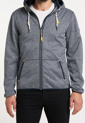 Muž ve šedé mikině na zip se žlutým jezdcem, šňůrkami se dřevěnými korálky a černých kalhotách stojí před bílým pozadím.