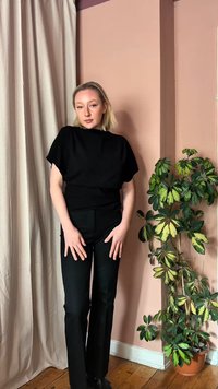 Joven con cabello rubio, vestida completamente de negro, de pie cerca de una cortina beige y una planta verde en maceta frente a una pared rosa apagado.