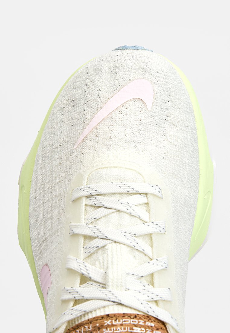 Zapatilla blanca con parte superior texturizada, swoosh de Nike en rosa, acentos grises, suela verde y cordones beige. Detalle notable de corcho en la lengüeta.