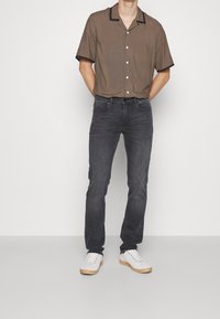 Korte mouwen bruin gestippeld shirt met zwarte accenten, gedragen met donkergrijze jeans en witte sneakers, staand op een effen achtergrond.