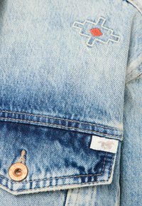 Jasnoniebieska kurtka dżinsowa z efektem gradientu, z haftowanym geometrycznym wzorem w kolorze czerwonym i białym. Mosiężne elementy metalowe i detale metki.