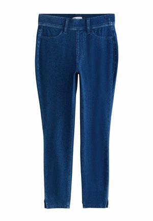 Femeie purtând un hanorac roșu supradimensionat cu un imprimeu colorat al unei scene dintr-o cafenea, cămașă albă cu guler, colanți bleumarin și balerini negri, stând în interior.