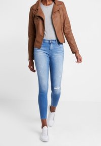 Bruin leren motorjack, grijze shirt, lichtblauwe versleten skinny jeans en witte sneakers. Het jack heeft een ritsluiting en schouderdetails.