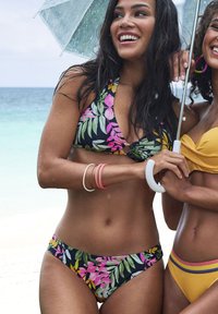 Dvije žene stoje u kupaćim kostimima uz plažu. Jedna nosi crni bikini s tropskim cvjetnim uzorkom; druga ima žuti top bez uzorka.