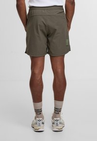 Pantalones cortos verde oliva con cinturilla elástica, bolsillos laterales y un pequeño detalle de parche. Llevados con calcetines beiges y zapatillas de running ligeras.