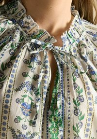 Blouse en tissu blanc avec des motifs floraux bleus et verts, encolure froncée avec un bord à volants, et une fermeture à nouer au col.