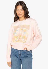 Rosa Sweatshirt mit einem bunten Grafikdruck, der Sonne, Auge und florale Motive zeigt. Rippbündchen und Saum. Kombiniert mit hellen Jeans.